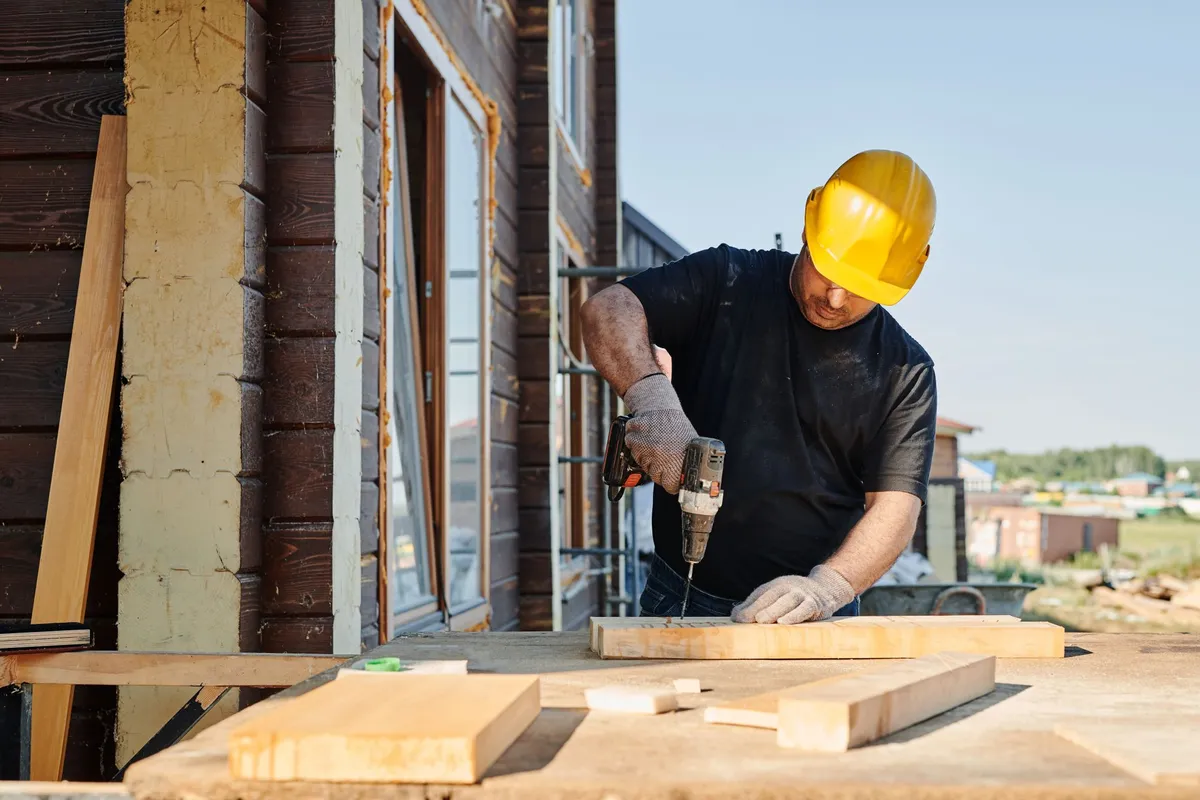 Construction worker in a yellow hard hat and black work shirt using a drill on timber outside an unfinished building