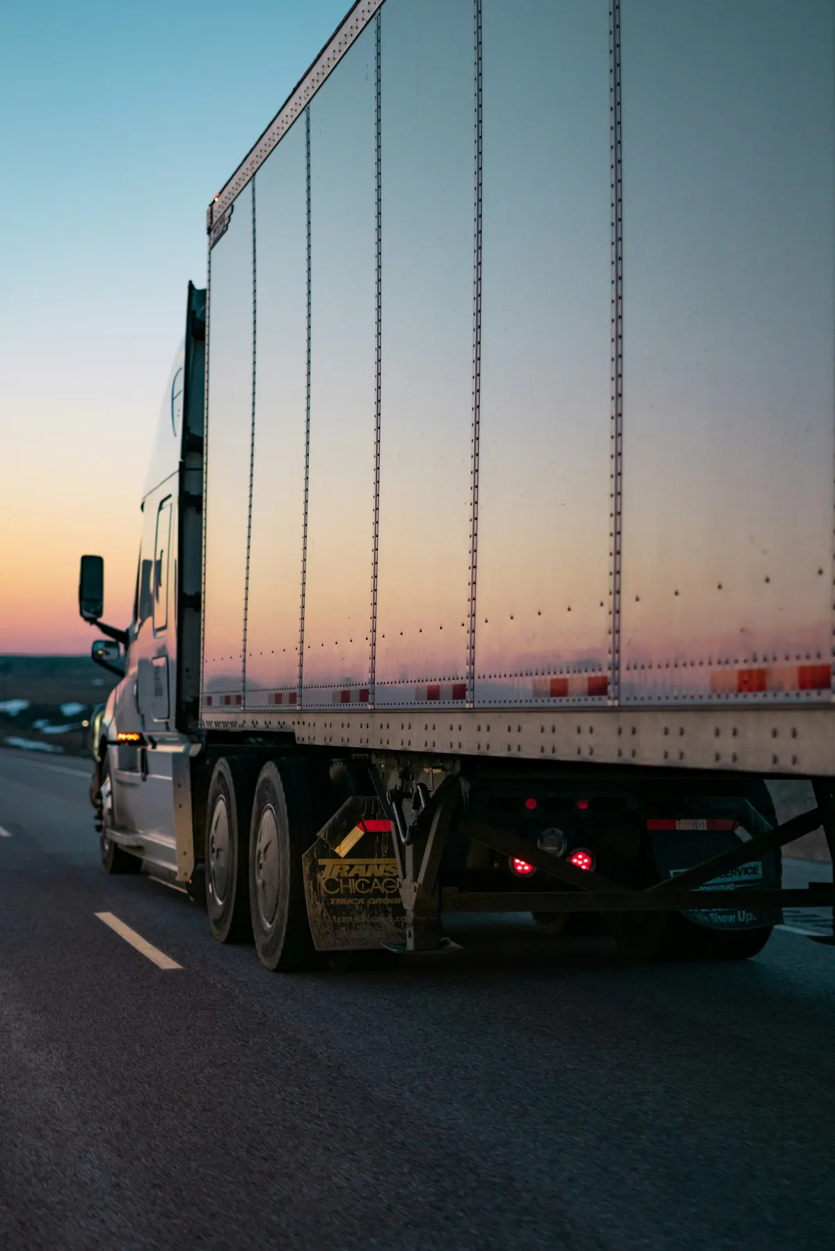 White tractor-trailer traveling on a highway at sunset, seen from behind.